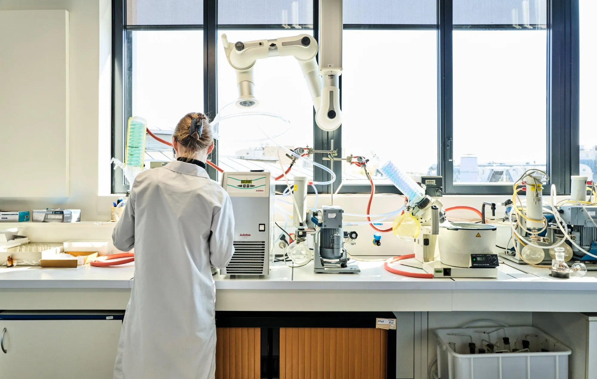 An engineering student in a lab coat works at a chemistry bench equipped with rotary evaporators, in front of a large bay window.